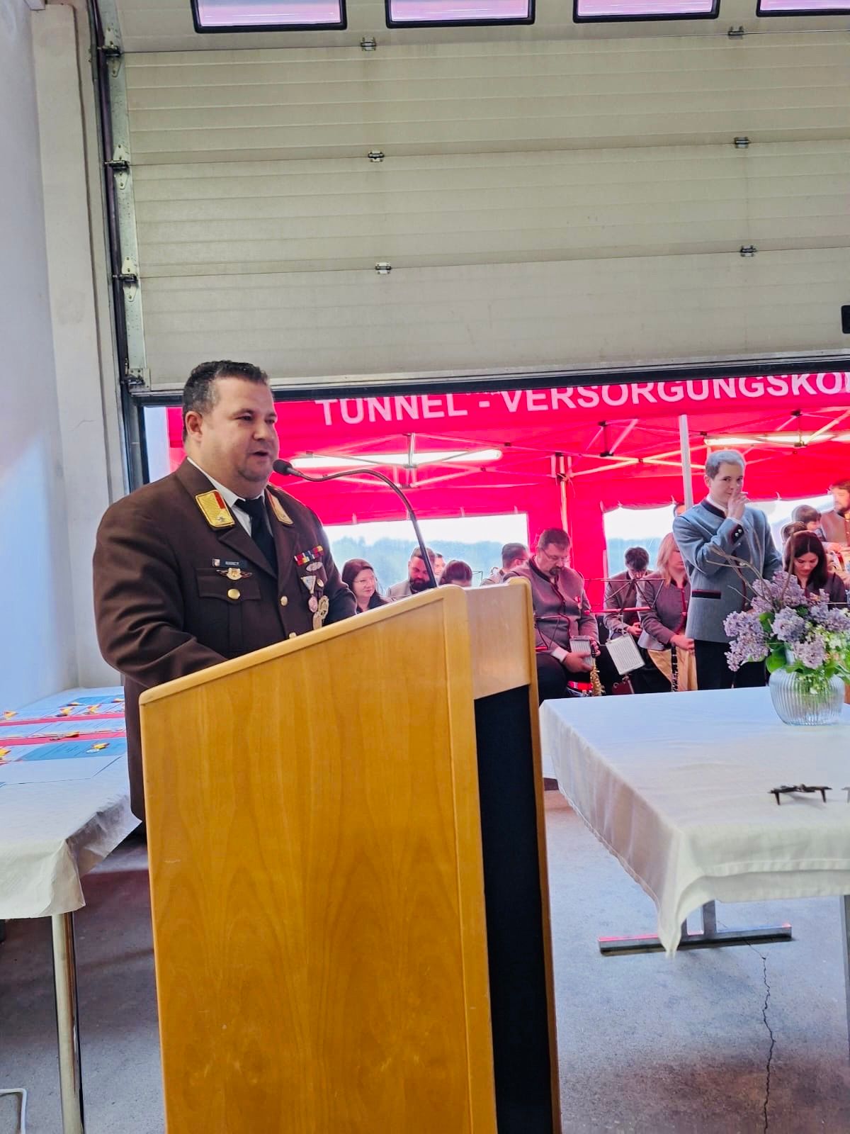 Ein Mann in Uniform steht an einem Rednerpult mit Mikrofon und spricht zu einem Publikum. Hinter ihm steht ein roter Banner mit der Aufschrift 'Tunnel-Versorgungskonzept'. Im Hintergrund sitzen und stehen Menschen, einige mit Blumen.