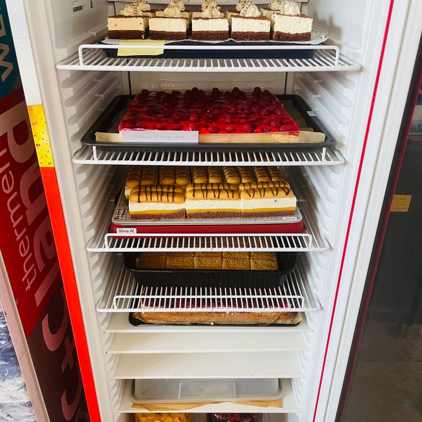 Ein Kühlschrank mit vier Regalen, gefüllt mit verschiedenen Desserts. Das oberste Regal hat Schokoladenkuchen, das zweite hat Erdbeertörtchen, das dritte hat gestreifte Kuchen und das unterste hat andere Desserts.
