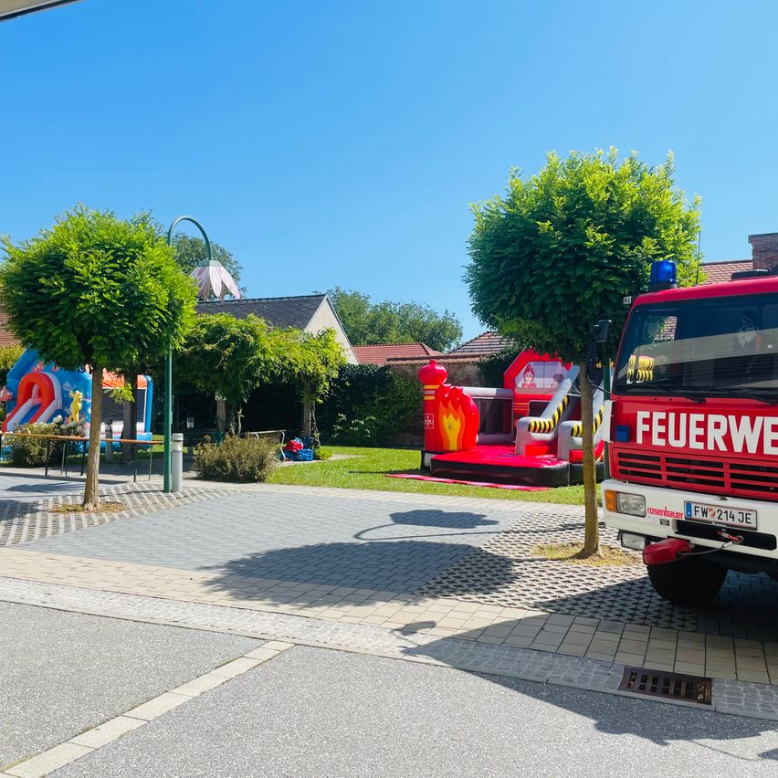 Ein Feuerwehrauto steht in einem Park mit einem Hüpfburg und Bäumen. Ein Gebäude mit Schornstein befindet sich hinter dem Feuerwehrauto. Der Himmel ist klar und blau.
