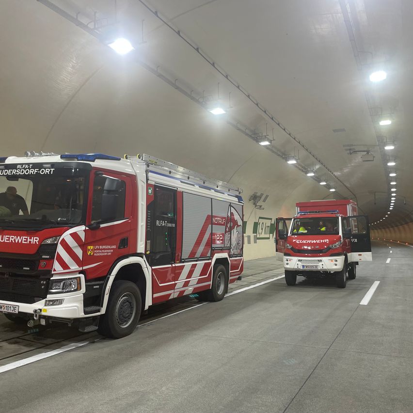 Zwei rote und weiße Feuerwehrfahrzeuge befinden sich in einem Tunnel, eines geparkt und das andere fährt aus. Beide Fahrzeuge haben Notsignale eingeschaltet. Der Tunnel ist mit weißen Deckenleuchten ausgestattet.