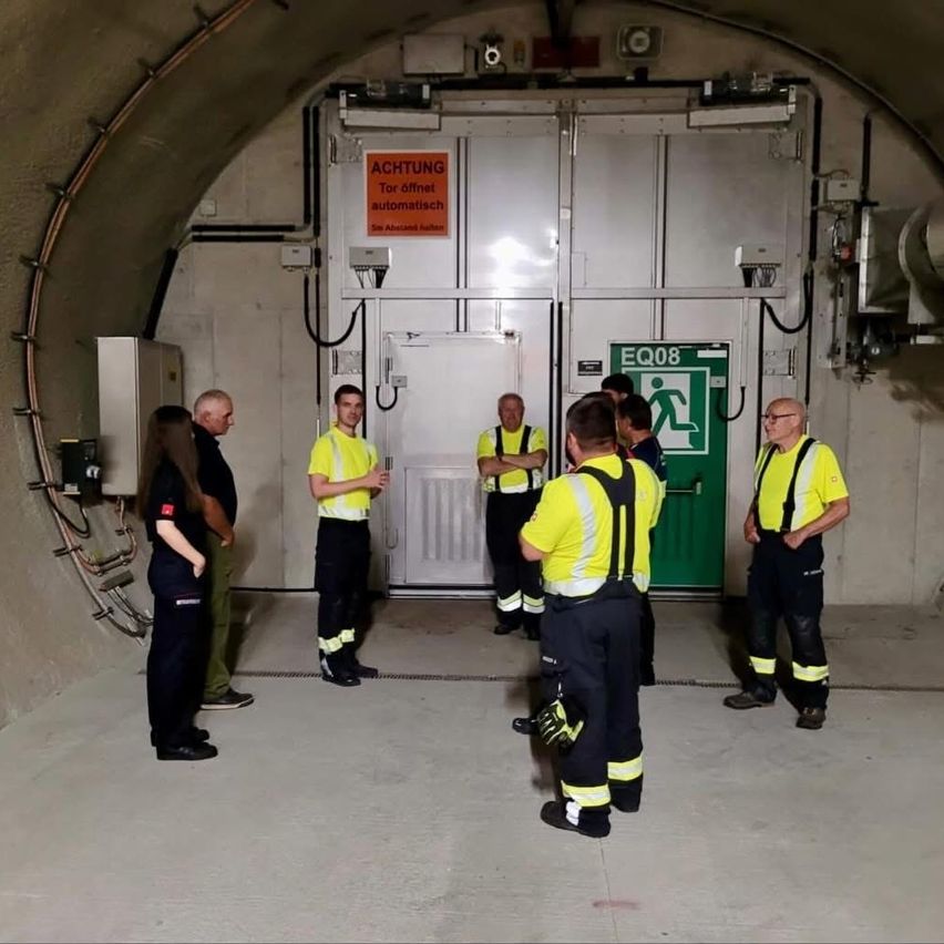 Eine Gruppe von Menschen in gelben Sicherheitsuniformen steht in einem Tunnel mit einer offenen Tür, die mit EQ08 beschriftet ist. Die Tür ist weiß mit einem grünen Notfallausgangsschild.
