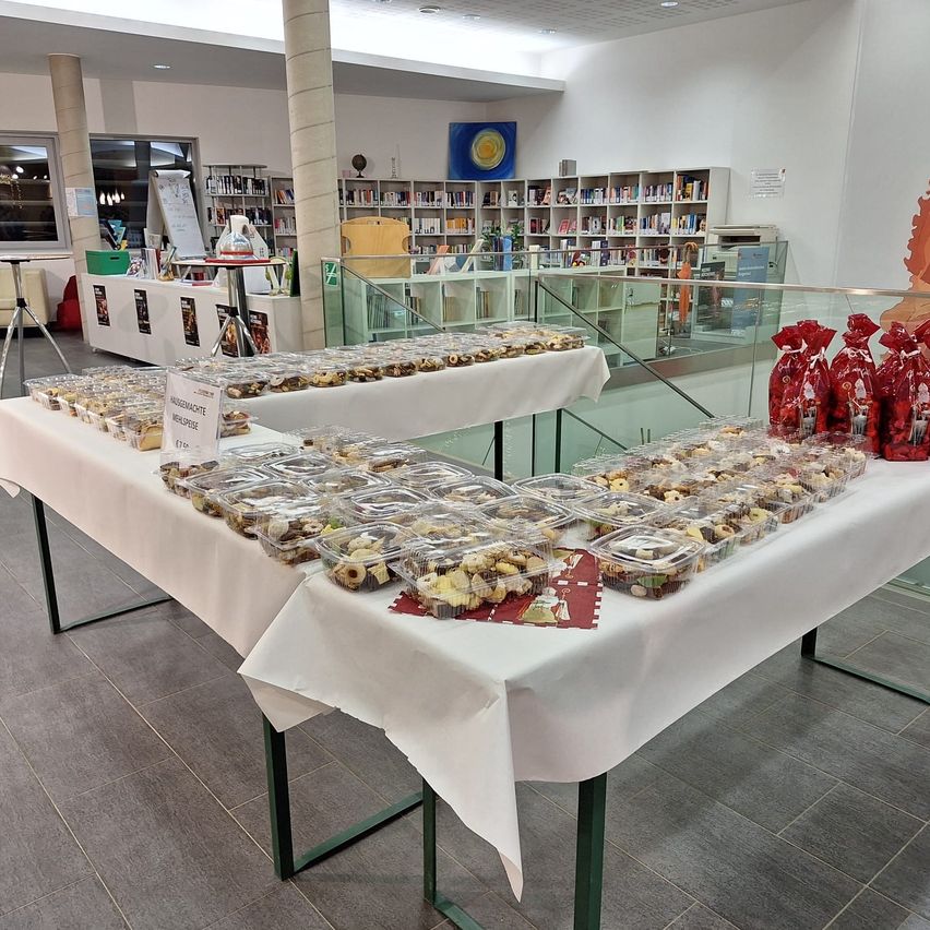 Eine Bibliothek mit einem Tisch voller verpackter Desserts, einschließlich Schokoladen, in durchsichtigen Behältern. Auf dem Tisch liegen auch rote Tüten. Der Raum hat Regale, die mit Büchern gefüllt sind.