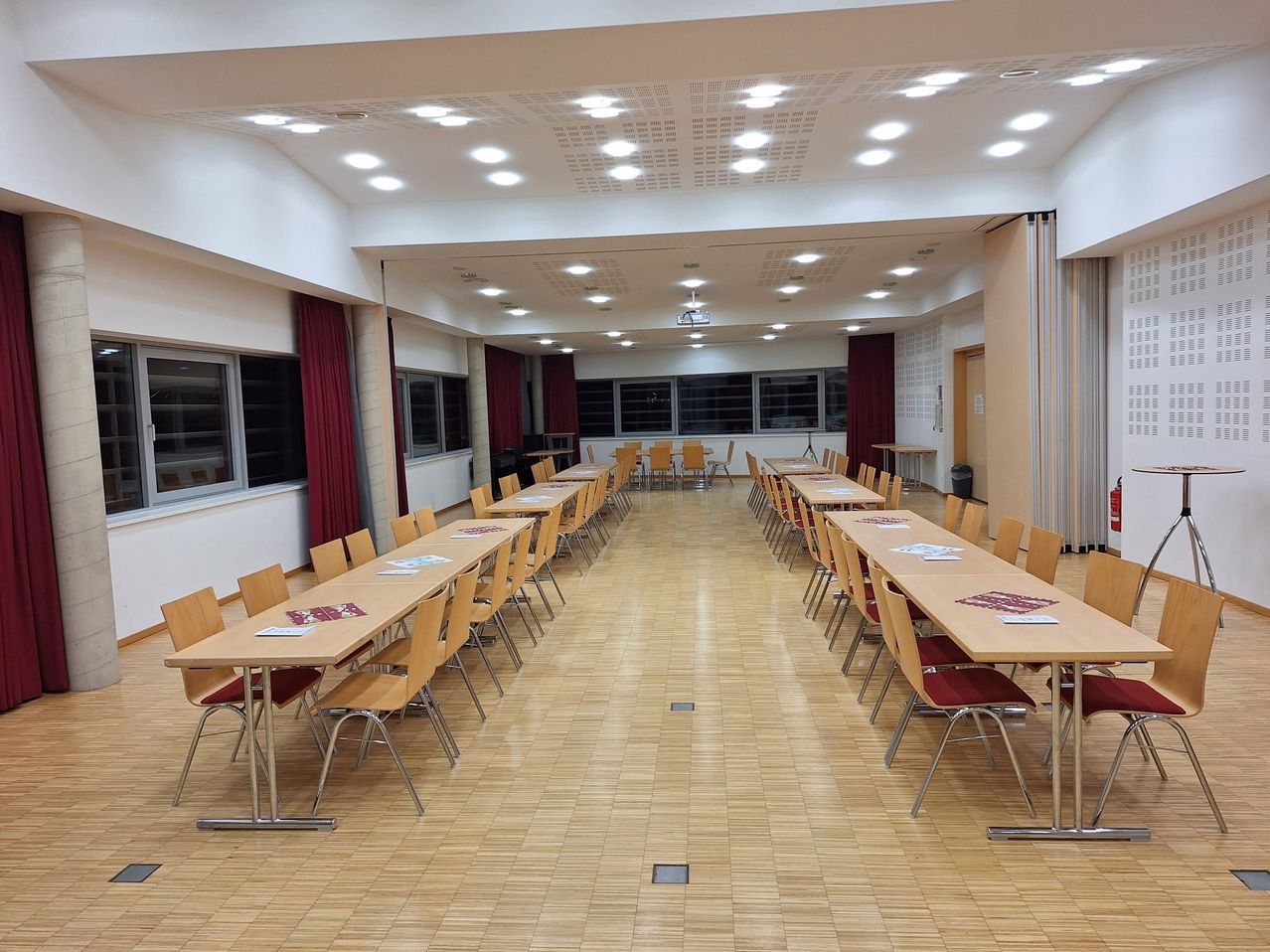 Ein leerer Konferenzraum mit Reihen von Holztischen und Stühlen, gefliestem Boden und geschlossenen Fenstern mit roten Vorhängen.