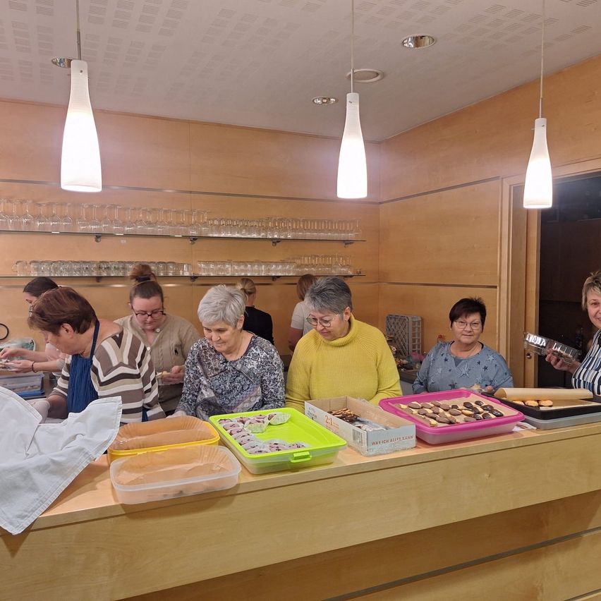 Eine Gruppe von Frauen bereitet in einer Küche mit Holzschränken, Gläserregalen und Lampen Essen zu. Auf dem Tresen stehen Tabletts und Schachteln mit Essen.