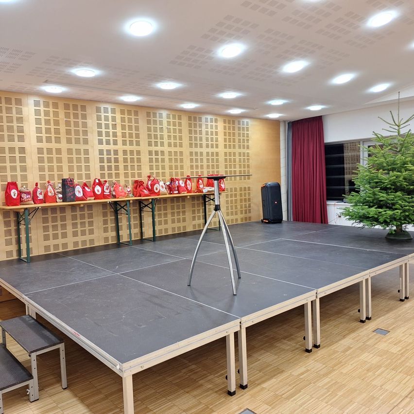 Ein Raum mit einer großen Bühne in der Mitte, Holzboden und einer Wand mit Aufbewahrungsboxen und einem Weihnachtsbaum. Die Bühne hat ein Stativ, und ein Lautsprecher steht an der Seite.