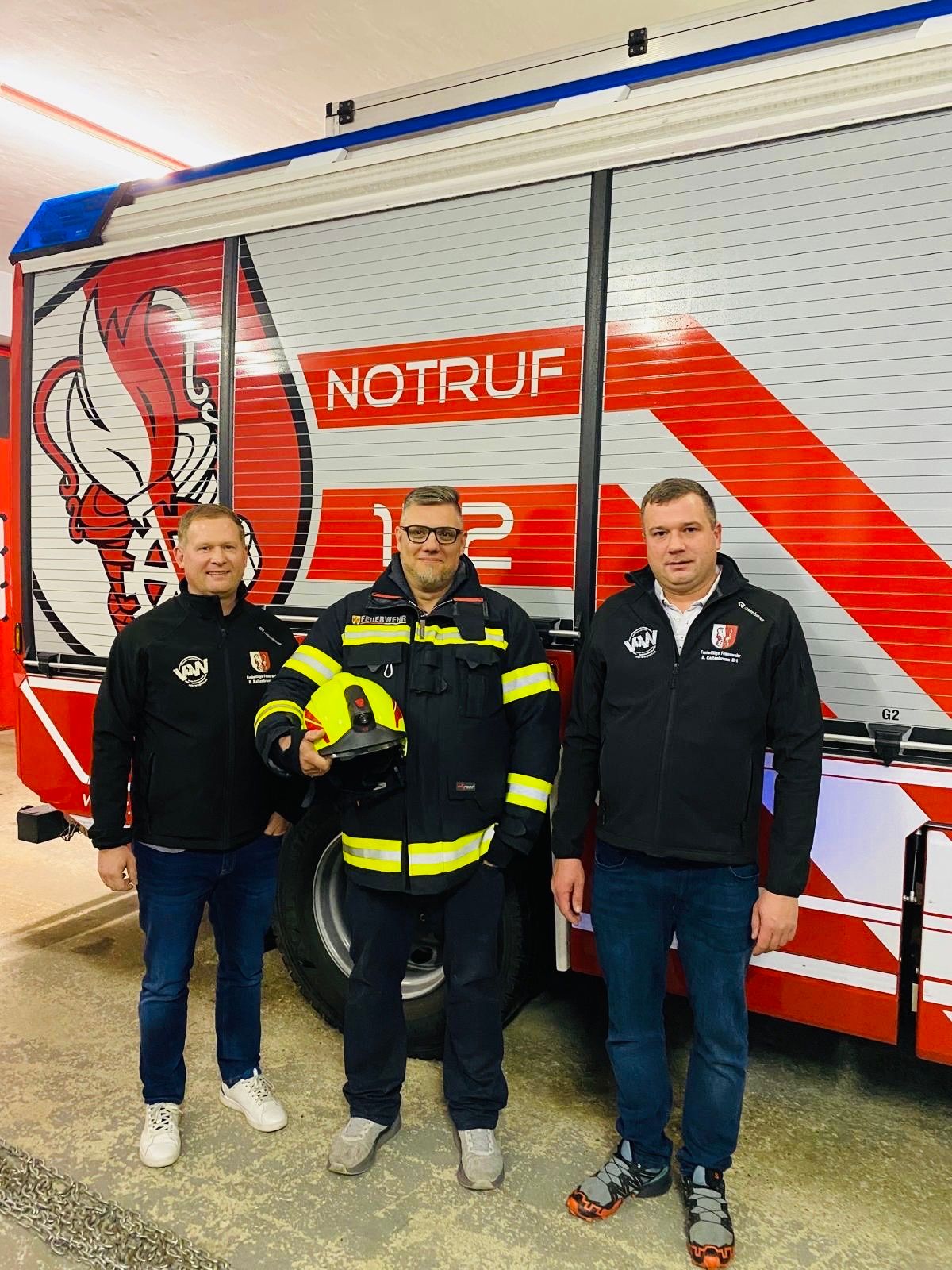 Three men stand in front of a fire truck labeled NOTRUF. The man in the middle holds a yellow helmet.