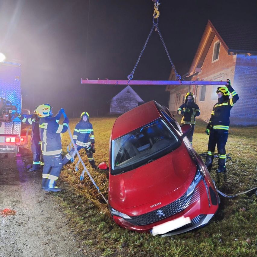 Ein rotes Auto ist auf der Straße umgekippt, Feuerwehrleute benutzen einen Kran, um es anzuheben. Die Szene ist nachts.