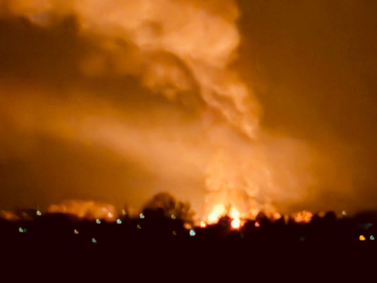 Ein großes Feuer in der Nacht mit aufsteigendem Rauch. Im Vordergrund sind Bäume und Lichter zu sehen.