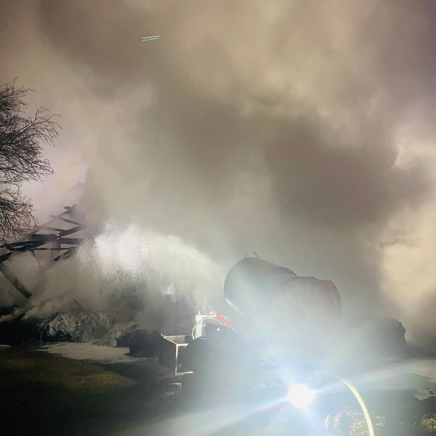 Feuerwehrleute löschen bei Nacht ein Feuer mit einem Wasserschlauch. Die Überreste eines Gebäudes und Rauch füllen die Luft.