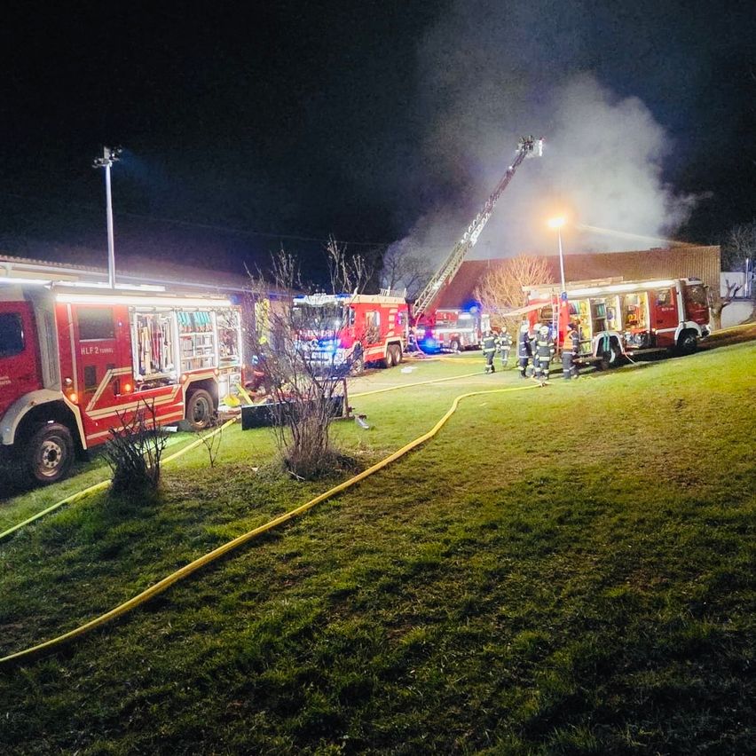 Mehrere Feuerwehrfahrzeuge sind auf einem Rasenfeld geparkt, mit aufsteigendem Rauch im Hintergrund. Eine Feuerwehrleiter ist auf das Gebäude ausgerichtet, wo Feuerwehrleute arbeiten.