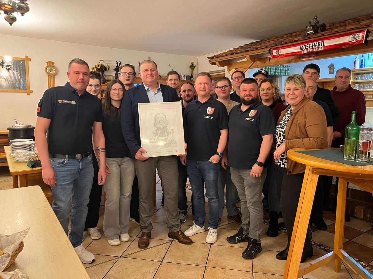 Eine Gruppe von Menschen posiert für ein Foto, wobei ein Mann in der Mitte ein gerahmtes Bild hält. Sie tragen passende dunkelblaue Poloshirts und Jeans. Der Raum hat geflieste Böden, einen Holztisch und ein Banner mit der Aufschrift 'MOST-HART'.