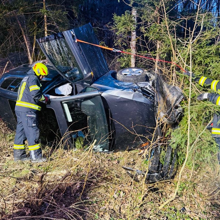 Feuerwehrleute retten ein Fahrzeug aus dem Gebüsch. Das Auto ist schwer beschädigt, mit offenem Motorhaube und abgerissenem Vorderrad.