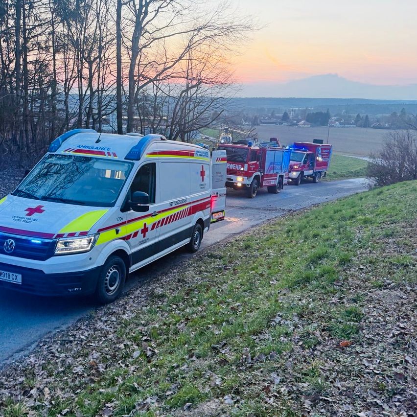 Ein Krankenwagen und Feuerwehrfahrzeuge sind am Straßenrand geparkt. Bäume säumen die Straße, und der Himmel ist eine Mischung aus Orange und Blau.