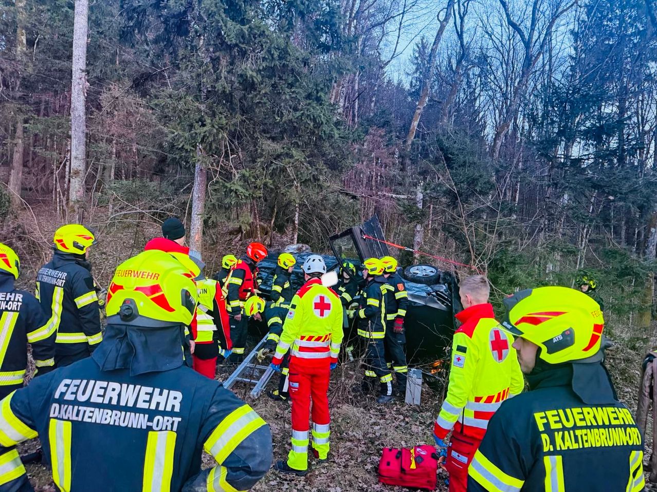 Rettungskräfte in gelben Helmen versammeln sich um ein beschädigtes Fahrzeug in einem bewaldeten Gebiet. Die Szene beinhaltet ein umgestürztes Auto, und die Einsatzkräfte kümmern sich um Personen.