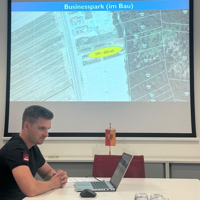 Ein Mann sitzt an einem Schreibtisch mit einem Laptop vor einer Projektionsleinwand, die eine Karte eines Gewerbeparks zeigt.