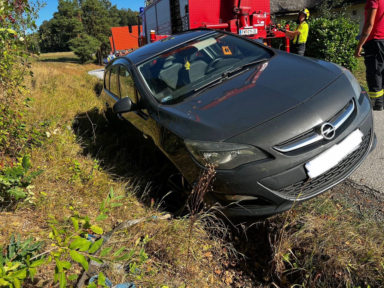 Ein schwarzer Opel Astra steht abseits der Straße in einem Grasfeld, mit einem Feuerwehrwagen in der Nähe. Eine Person in einer gelben Weste steht neben dem Feuerwehrwagen.