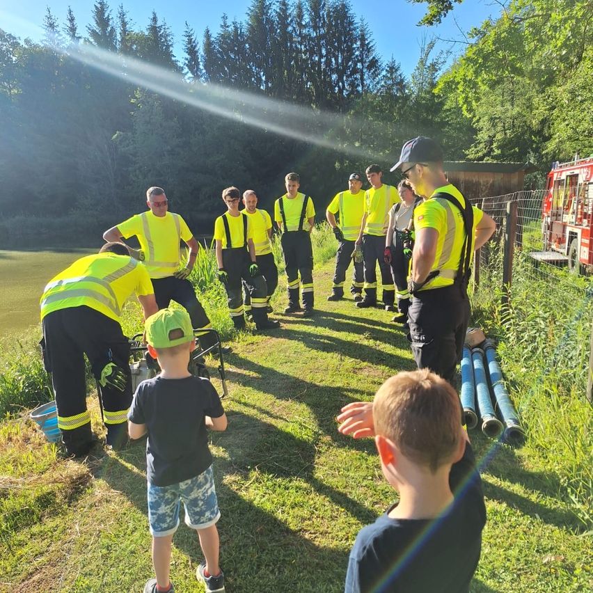 Eine Gruppe von Feuerwehrleuten in leuchtend gelben Westen steht in der Nähe eines Sees, ein Feuerwehrauto ist in der Nähe geparkt. Zwei Kinder beobachten sie, eines hält einen Eimer.