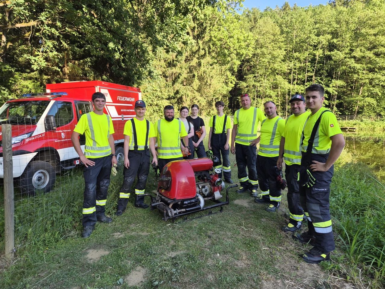 Eine Gruppe von Feuerwehrleuten in Warnwesten posiert für ein Foto im Freien mit einem Feuerwehrwagen und einem roten Maschinenfahrzeug im Hintergrund.