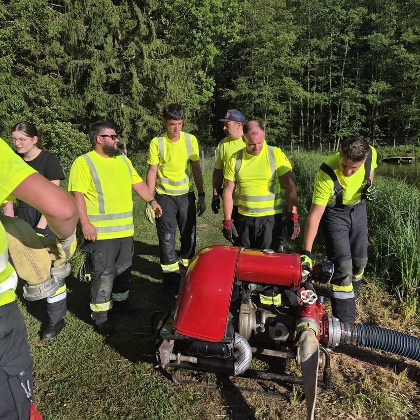 Eine Gruppe von Feuerwehrleuten in leuchtend gelben Uniformen steht in der Nähe einer roten Pumpe auf einem Rasengebiet nahe einem Wald.