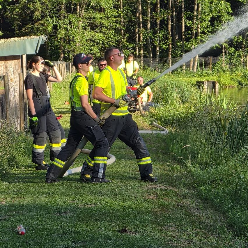 Feuerwehrleute üben mit einem Wasserschlauch auf einer Wiese in der Nähe eines Teichs. Sie tragen gelbe Uniformen und Helme. Eine Frau in Schwarz beobachtet von links.