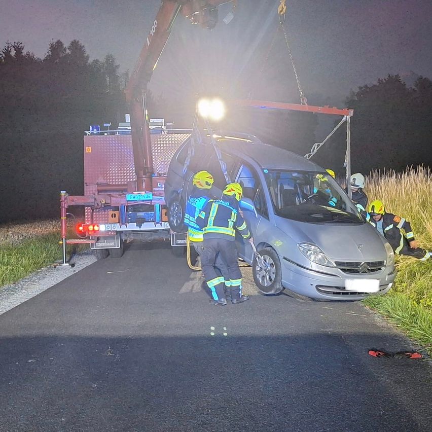 Ein Kran hebt ein beschädigtes Auto auf einer Straße bei Nacht. Vier Personen in Warnwesten und Helmen stehen darum.