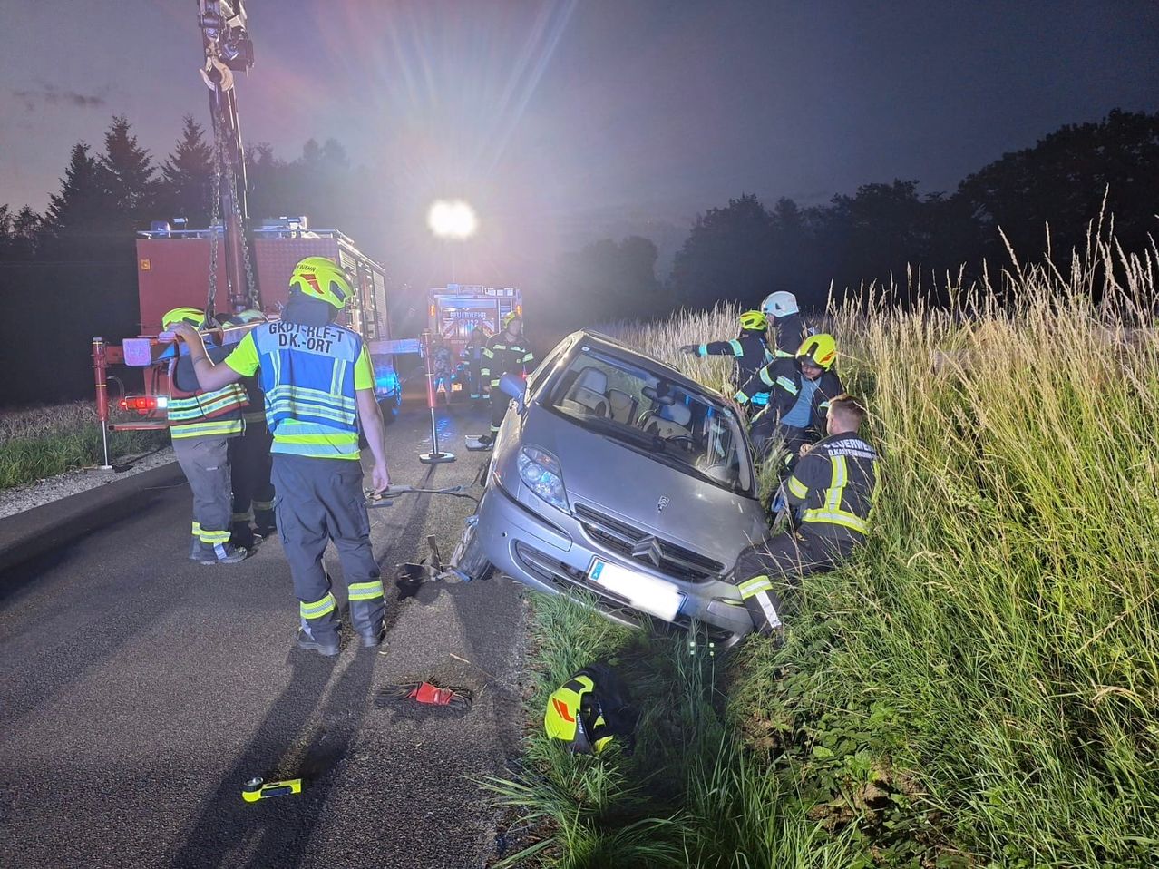 Rettungskräfte helfen bei einem beschädigten Auto am Straßenrand, Feuerwehrleute in Helmen und Warnwesten in der Nähe.