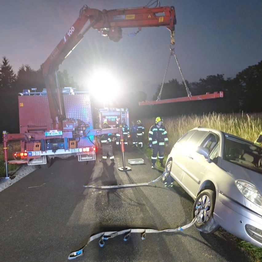 Feuerwehrleute benutzen einen Kran, um ein demoliertes Auto nachts von der Straße zu heben. Das Auto ist mit Seilen gesichert und von Rettungsfahrzeugen umgeben.