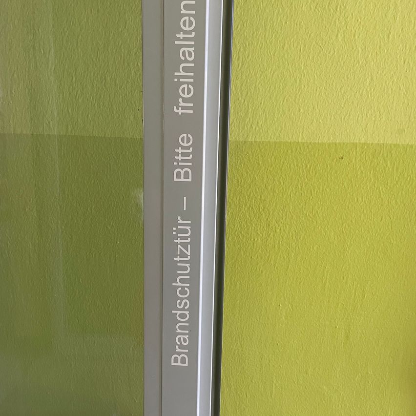 Eine Glastür mit einer gelben Wand im Hintergrund. Die Tür hat einen silbernen Griff und einen weißen Aufkleber mit dem Text 'Brandschutztür - Bitte freihalten'.