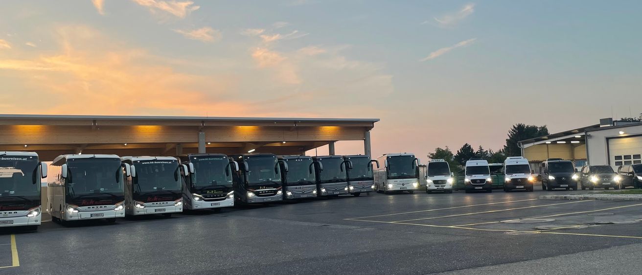 Mehrere Busse sind unter einem Schutzdach mit einem Sonnenuntergangshimmel darüber geparkt. Die Fahrzeuge sind aufgereiht, und einige haben Scheinwerfer an.