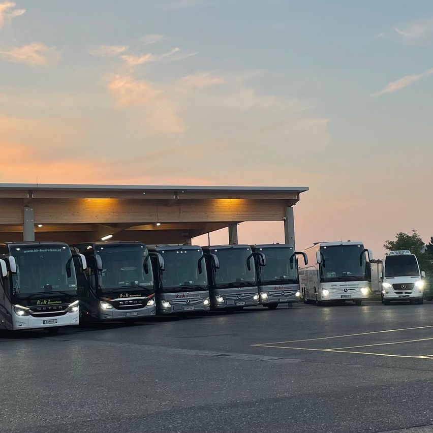 Mehrere Busse sind unter einem Schutzdach mit einem Sonnenuntergangshimmel darüber geparkt. Die Fahrzeuge sind aufgereiht, und einige haben Scheinwerfer an.