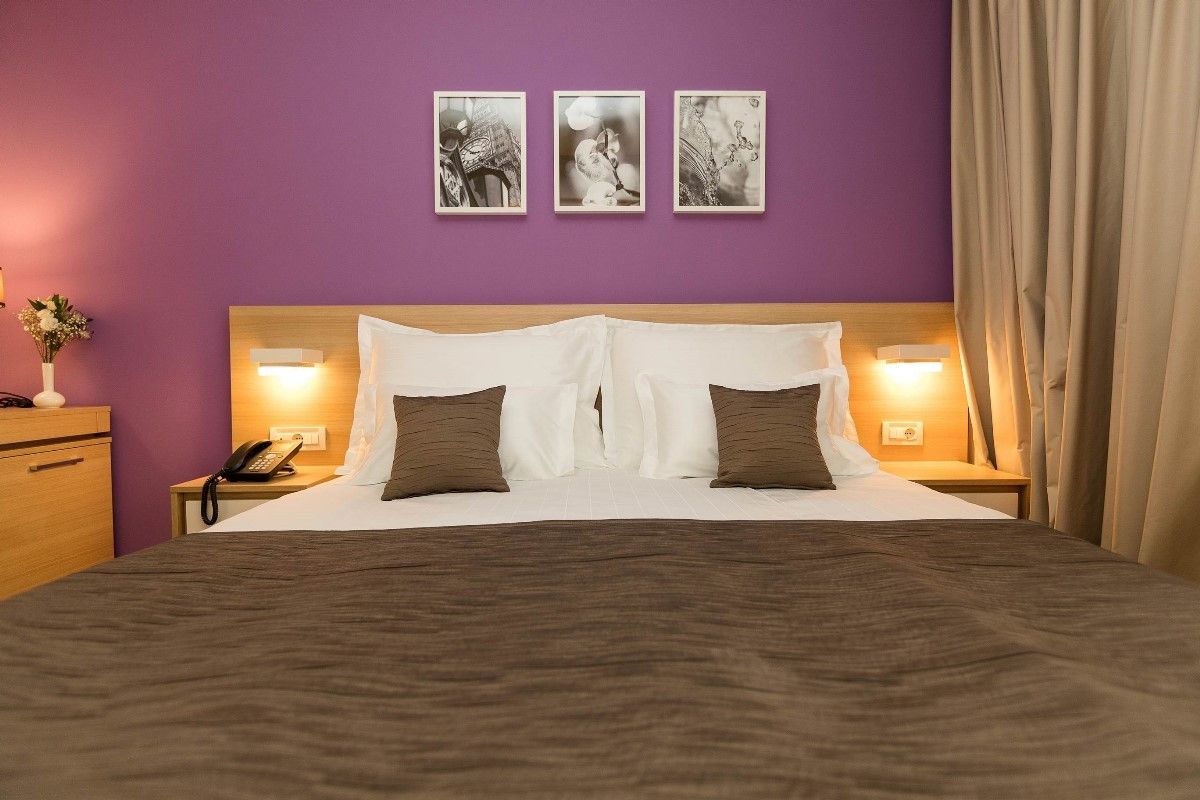 Ein Schlafzimmer mit einem großen Bett und lila Wänden, mit drei schwarz-weiß gerahmten Bildern darüber. Das Bett hat weiße Kissen und braune Kissen mit einer braunen Decke.