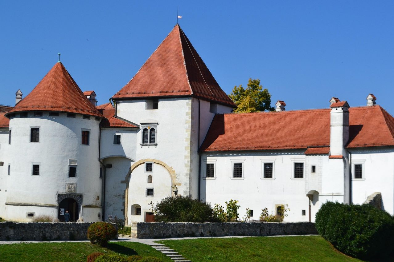 A large white castle with red roofs and several windows, under a clear blue sky, surrounded by greenery and a stone wall.