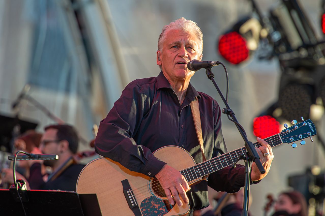 Ein älterer Mann mit weißen Haaren singt in ein Mikrofon, während er eine Gitarre hält. Im Hintergrund spielt ein Cellist.