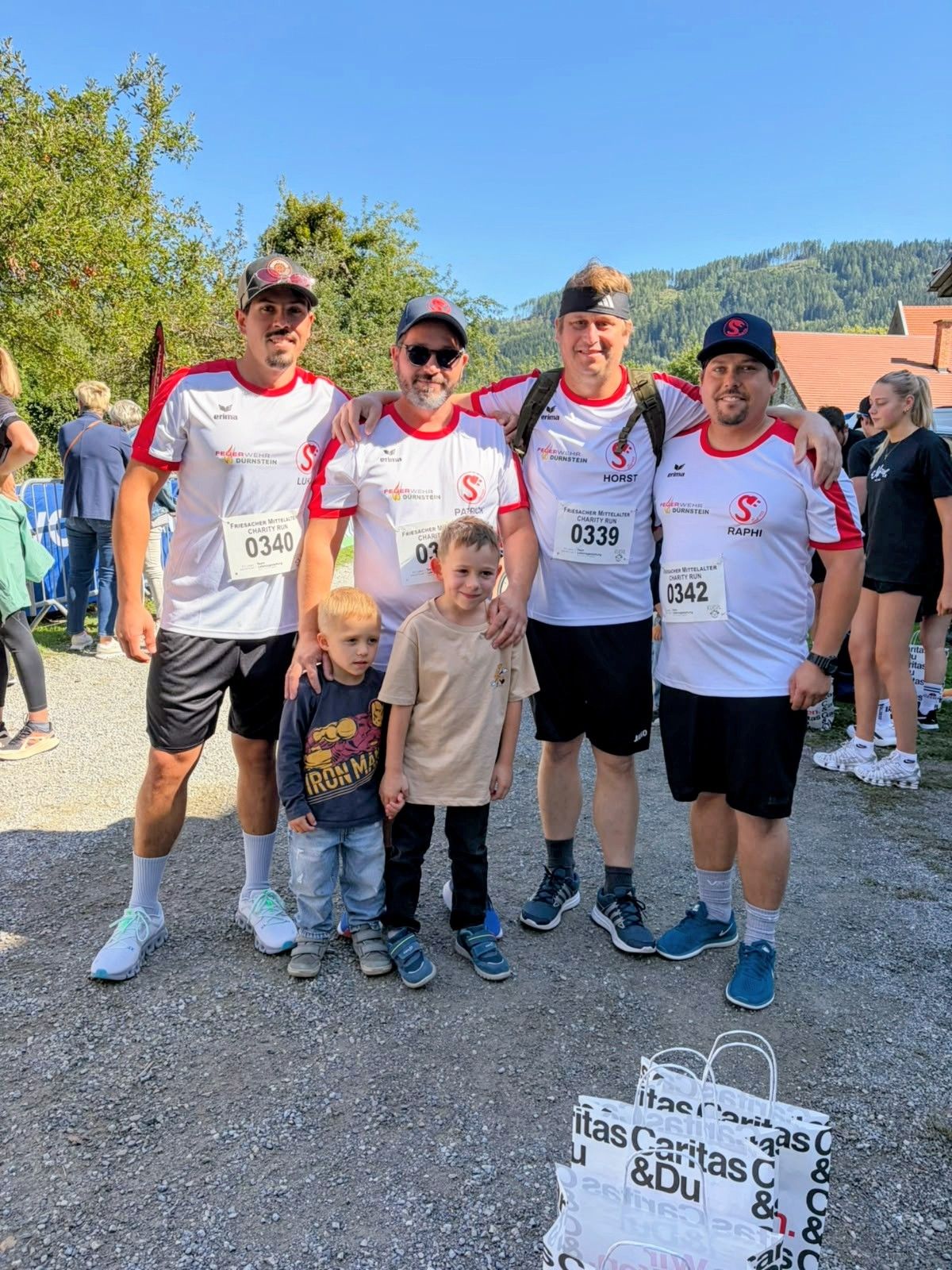 Vier Männer und zwei Jungen posieren für ein Foto bei einem Wohltätigkeitslauf. Sie tragen passende Shirts mit Nummern und stehen auf einem Schotterweg.