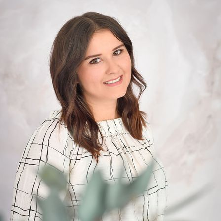 Eine Frau mit braunen Haaren und einem weißen Hemd mit schwarzen Linien posiert für ein Foto und lächelt, mit einer Pflanze im Vordergrund.