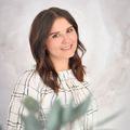Eine Frau mit braunen Haaren und einem weißen Hemd mit schwarzen Linien posiert für ein Foto und lächelt, mit einer Pflanze im Vordergrund.