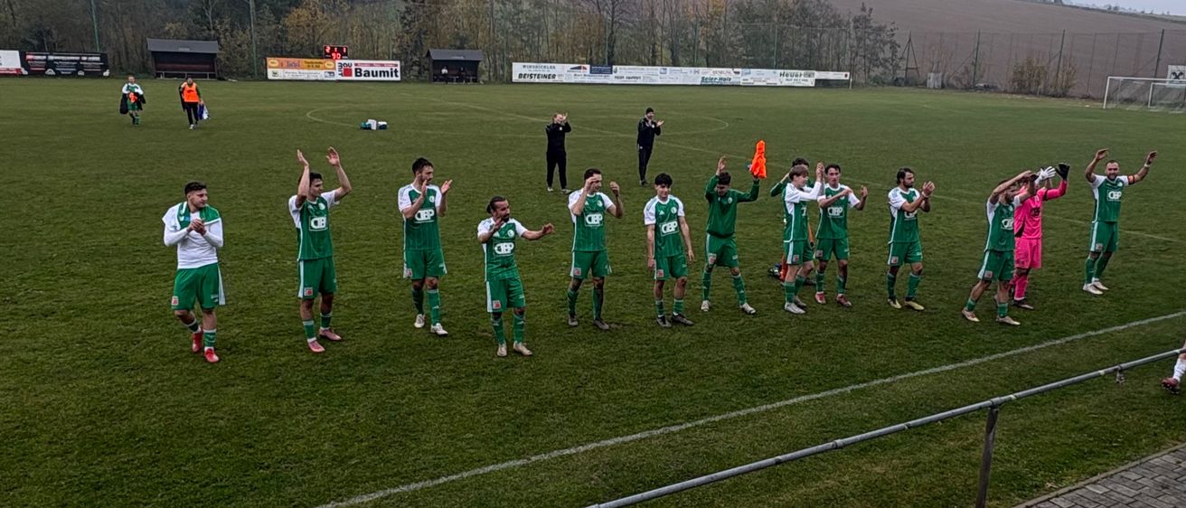 Fußballspieler in grünen Uniformen stellen sich auf dem Feld auf, möglicherweise zum Feiern. Das Feld ist von Bäumen und einer Umzäunung umgeben.