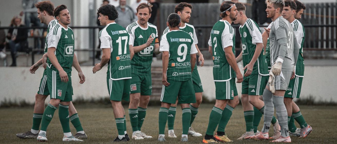 Eine Fußballmannschaft in grünen und weißen Trikots steht auf einem Spielfeld. Die Spieler schauen in eine Richtung, vielleicht diskutieren sie über eine Spielstrategie. Im Hintergrund befindet sich ein Gebäude mit Text an der Wand.