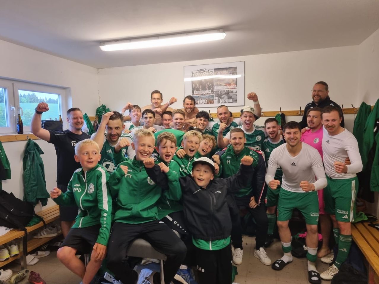 Eine Gruppe von Fußballspielern und Trainern posiert für ein Foto im Umkleideraum. Die Spieler tragen grüne und weiße Trikots, während die Trainer schwarze und rosa Hemden tragen. Ein Banner hängt an der Wand.