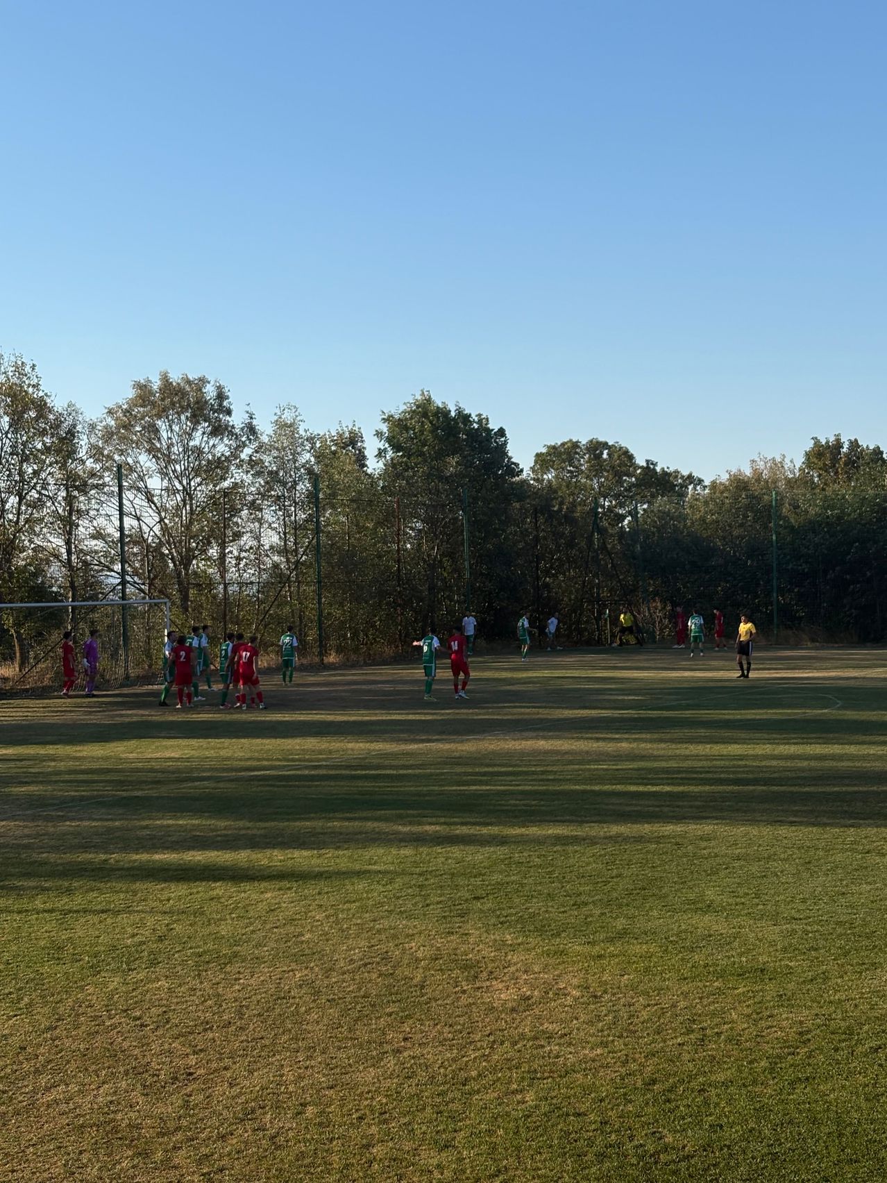 A soccer match is underway on a grassy field with players in red and green uniforms. The field is surrounded by trees and a fence.
