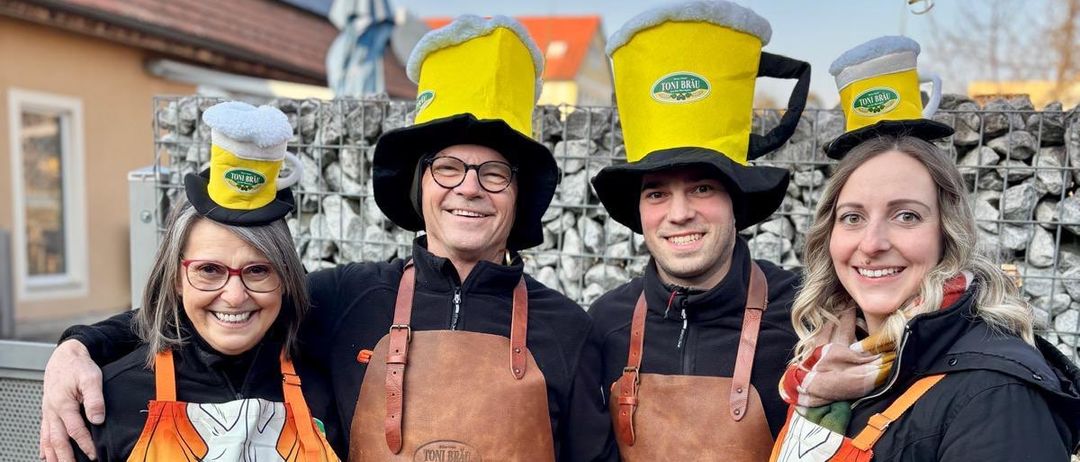 Vier Menschen in Schürzen und Hüten stehen lächelnd vor einer Steinmauer. Zwei Männer tragen braune Schürzen, zwei Frauen orangene Schürzen. Ein gelber Ballon schwebt darüber.