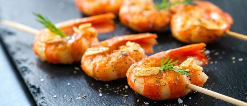 Eine Nahaufnahme von gegrillten Garnelen auf Spießen, garniert mit Kräutern und Gewürzen, serviert auf einer schwarzen Schieferplatte mit einem Salat im Hintergrund.