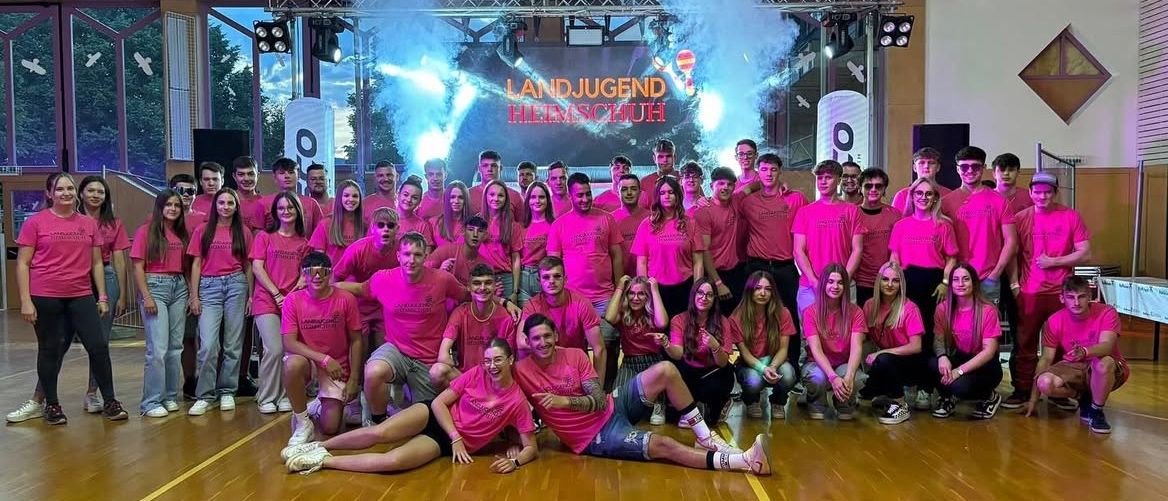 Eine Gruppe von Menschen in rosa Shirts und mit Lächeln posiert für ein Foto auf einer Bühne mit rosa Rauch im Hintergrund.