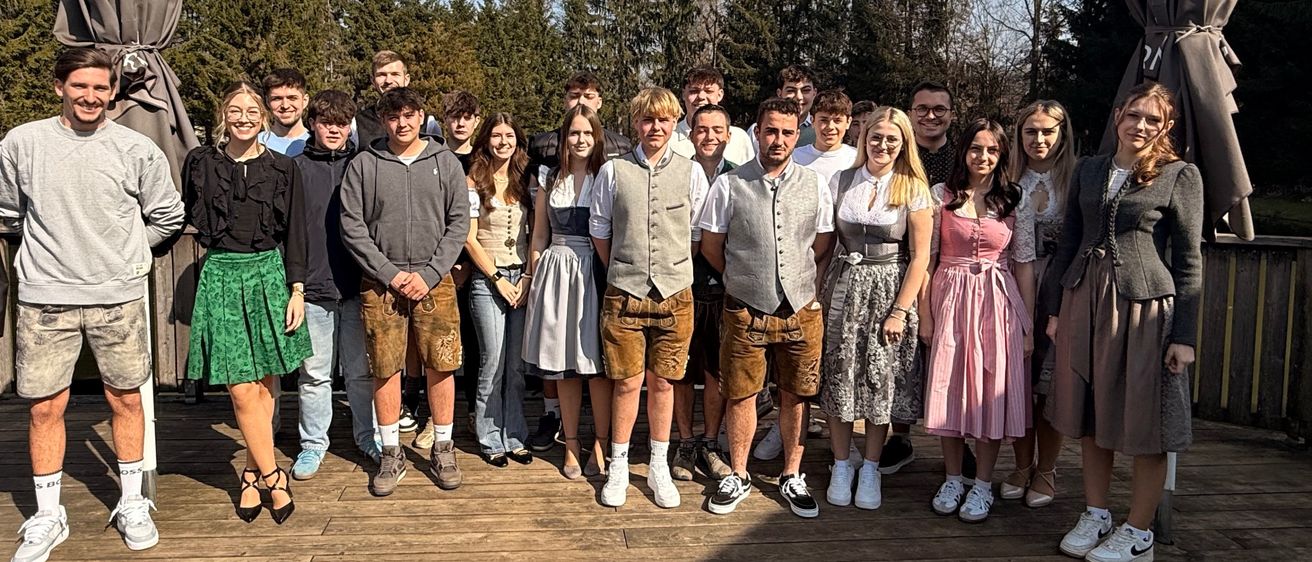 Eine Gruppe von Männern und Frauen steht zusammen auf einer Holzterrasse mit Bäumen im Hintergrund. Sie tragen Freizeit- und traditionelle Kleidung.