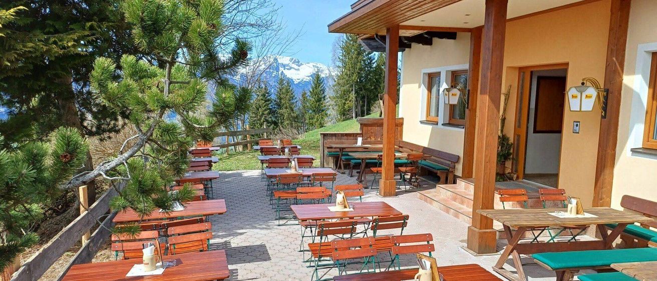 Terrassenbereich mit Holztischen und Stühlen vor der Kulisse von Bergen und Bäumen. Das Restaurant verfügt über Holzbänke und Tische unter einem überdachten Vordach.