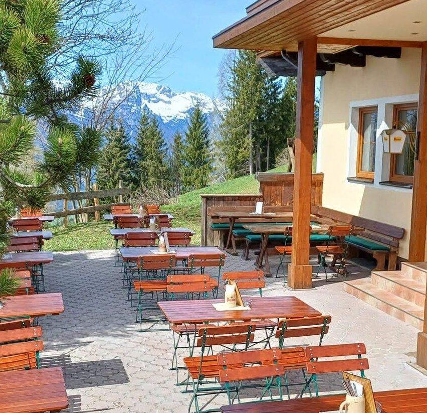 Terrassenbereich mit Holztischen und Stühlen vor der Kulisse von Bergen und Bäumen. Das Restaurant verfügt über Holzbänke und Tische unter einem überdachten Vordach.