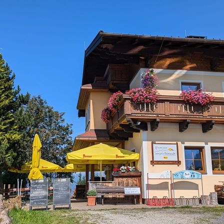 Ein rustikales Gebäude mit einem Balkon, geschmückt mit rosa Blumen, gelben Sonnenschirmen und einem Schild draußen. Es hat eine Bank, Topfpflanzen und Fahrräder davor.