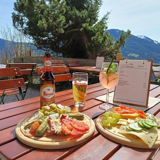 Ein Holztisch im Freien mit zwei Tellern Essen, einer Flasche Bier und einem Glas Getränk. Der Tisch ist von Holzstühlen umgeben. In der Ferne befindet sich eine Bergkette.