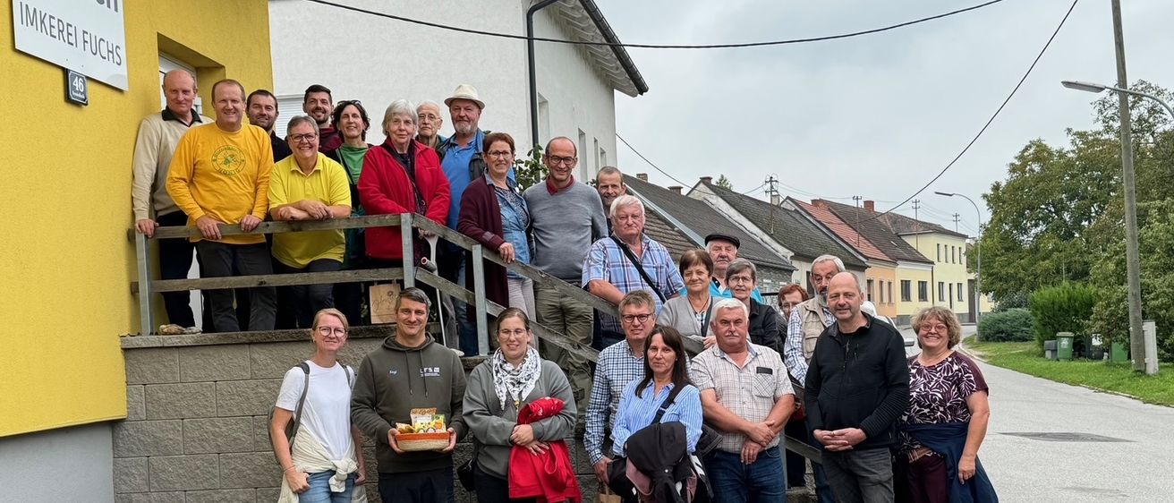Eine Gruppe von Menschen posiert für ein Foto vor einem Gebäude mit gelben Wänden und einer Holztreppe. Einige tragen Hüte, Brillen und Turnschuhe. Sie scheinen auf einer Reise zu sein.