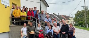 Eine Gruppe von Menschen posiert für ein Foto vor einem Gebäude mit gelben Wänden und einer Holztreppe. Einige tragen Hüte, Brillen und Turnschuhe. Sie scheinen auf einer Reise zu sein.